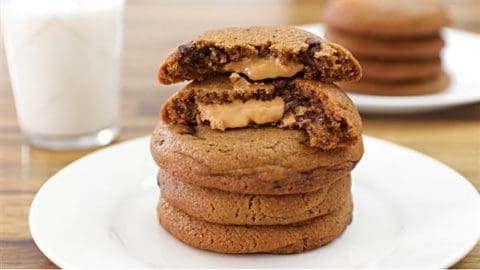 A stack of chocolate chip cookies is on a white plate. The top cookie is broken in half, revealing a creamy peanut butter filling. A glass of milk and another plate with more cookies are visible in the background.