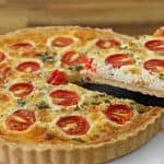 A quiche with a golden-brown crust sits on a white plate. The filling is topped with halved cherry tomatoes and sprinkled with herbs. A cut slice is being lifted out with a serving utensil, revealing the creamy interior. The background features a wooden surface.