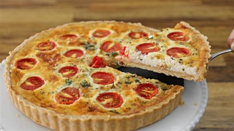 A quiche with a golden-brown crust sits on a white plate. The filling is topped with halved cherry tomatoes and sprinkled with herbs. A cut slice is being lifted out with a serving utensil, revealing the creamy interior. The background features a wooden surface.