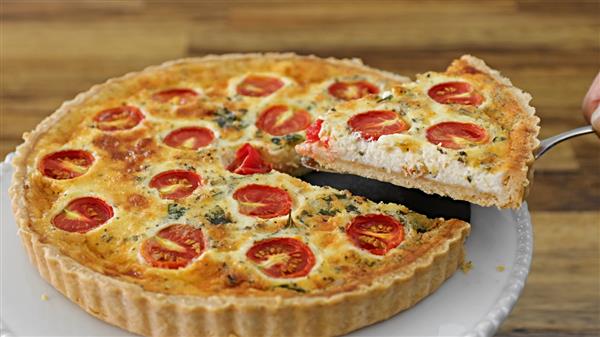 A quiche with a golden-brown crust sits on a white plate. The filling is topped with halved cherry tomatoes and sprinkled with herbs. A cut slice is being lifted out with a serving utensil, revealing the creamy interior. The background features a wooden surface.