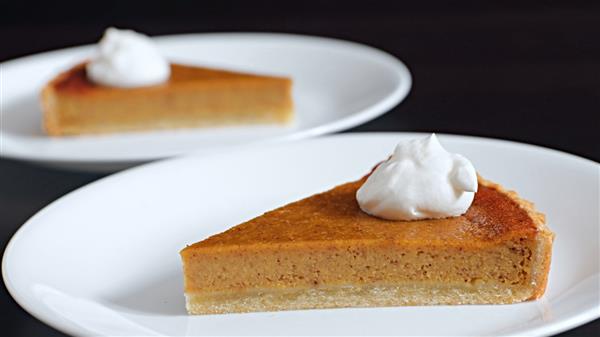 Two slices of pumpkin pie each topped with a dollop of whipped cream, served on white plates. The dessert features a smooth, creamy filling and a crisp, golden-brown crust. The background is dark, with the focus on the pie slices.