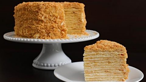 A multi-layered honey cake is displayed on a white pedestal cake stand against a dark background. A separate slice of the cake sits on a white plate in the foreground, showing its numerous thin layers and golden, crumb-coated exterior.