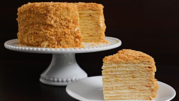 A multi-layered honey cake is displayed on a white pedestal cake stand against a dark background. A separate slice of the cake sits on a white plate in the foreground, showing its numerous thin layers and golden, crumb-coated exterior.