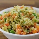 A white bowl filled with a colorful vegetable couscous salad, featuring fluffy couscous grains mixed with green peas, diced orange carrots, and green herbs. The bowl is set on a wooden surface.