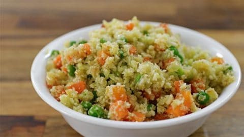 A white bowl filled with a colorful vegetable couscous salad, featuring fluffy couscous grains mixed with green peas, diced orange carrots, and green herbs. The bowl is set on a wooden surface.