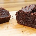 A loaf of chocolate chip banana bread lies on a wooden surface. One slice is cut and positioned next to the loaf, showing the moist, rich chocolate interior dotted with chocolate chips. The background is softly blurred.