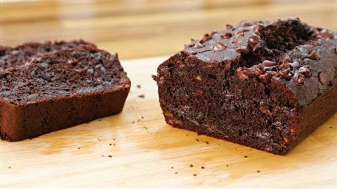 A loaf of chocolate chip banana bread lies on a wooden surface. One slice is cut and positioned next to the loaf, showing the moist, rich chocolate interior dotted with chocolate chips. The background is softly blurred.