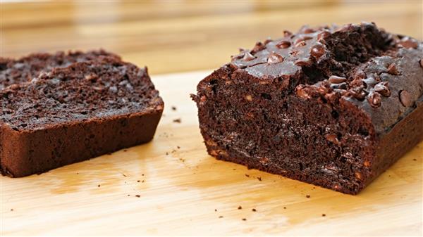 A loaf of chocolate chip banana bread lies on a wooden surface. One slice is cut and positioned next to the loaf, showing the moist, rich chocolate interior dotted with chocolate chips. The background is softly blurred.