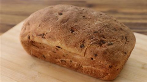 A freshly baked loaf of bread sits on a wooden surface. The bread has a golden-brown crust with visible flecks of what appears to be herbs or spices. The texture looks rustic and hearty, with a slightly domed top.