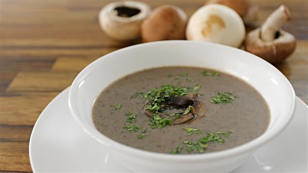 A white bowl filled with creamy mushroom soup garnished with fresh parsley and sliced mushrooms is placed on a matching white plate. In the background, various whole mushrooms are scattered on a wooden surface.