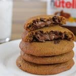 A stack of Nutella-filled cookies sits on a white plate, with the top cookie broken in half to reveal a gooey chocolate hazelnut filling. In the background, there is a glass of milk and a jar of Nutella.