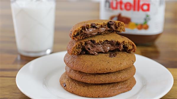 A stack of Nutella-filled cookies sits on a white plate, with the top cookie broken in half to reveal a gooey chocolate hazelnut filling. In the background, there is a glass of milk and a jar of Nutella.