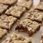 A tray of oatmeal chocolate squares arranged in rows on a parchment paper. Each square has a layer of melted chocolate topped with a generous sprinkling of oatmeal. One square in the front row has a bite taken out of it, revealing a dense texture inside.