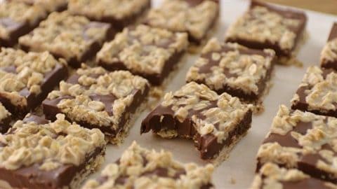 A tray of oatmeal chocolate squares arranged in rows on a parchment paper. Each square has a layer of melted chocolate topped with a generous sprinkling of oatmeal. One square in the front row has a bite taken out of it, revealing a dense texture inside.