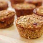 Close-up of freshly baked oatmeal muffins with a golden-brown crust, arranged on a light wooden surface. The muffins contain visible oats and appear moist and textured, suggesting a warm and homemade feel. Several muffins are in focus, while others are blurred in the background.