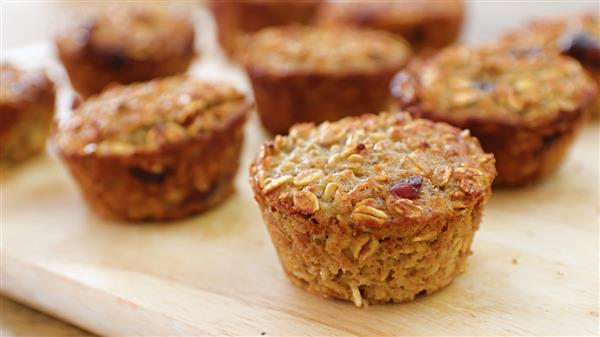 Close-up of freshly baked oatmeal muffins with a golden-brown crust, arranged on a light wooden surface. The muffins contain visible oats and appear moist and textured, suggesting a warm and homemade feel. Several muffins are in focus, while others are blurred in the background.