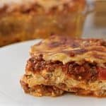 A slice of lasagna with layers of pasta, meat sauce, and melted cheese is served on a white plate. In the background, a glass baking dish with more lasagna is visible on a wooden table.