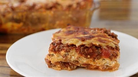 A slice of lasagna with layers of pasta, meat sauce, and melted cheese is served on a white plate. In the background, a glass baking dish with more lasagna is visible on a wooden table.