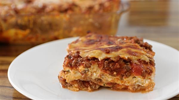 A slice of lasagna with layers of pasta, meat sauce, and melted cheese is served on a white plate. In the background, a glass baking dish with more lasagna is visible on a wooden table.