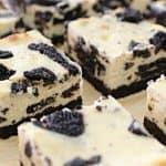 Squares of cookies and cream cheesecake are displayed, each featuring a creamy vanilla base mixed with chunks of chocolate sandwich cookies on a chocolate cookie crust. The surface is dotted with additional cookie pieces, creating a visually appealing dessert.