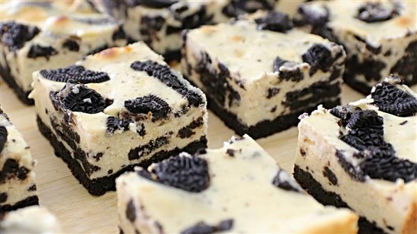 Squares of cookies and cream cheesecake are displayed, each featuring a creamy vanilla base mixed with chunks of chocolate sandwich cookies on a chocolate cookie crust. The surface is dotted with additional cookie pieces, creating a visually appealing dessert.