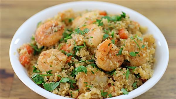 A white bowl filled with a mixture of shrimp, quinoa, diced vegetables, and garnished with chopped parsley, set on a wooden surface. The colorful dish highlights the shrimp and fresh herbs.