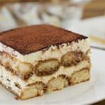 A slice of tiramisu is served on a white plate. The dessert features visible layers of coffee-soaked ladyfingers and mascarpone cream, topped with a dusting of cocoa powder. Another slice of tiramisu and the dessert tray are blurred in the background.