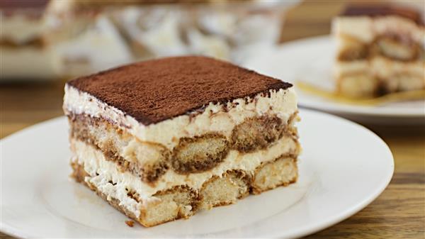 A slice of tiramisu is served on a white plate. The dessert features visible layers of coffee-soaked ladyfingers and mascarpone cream, topped with a dusting of cocoa powder. Another slice of tiramisu and the dessert tray are blurred in the background.