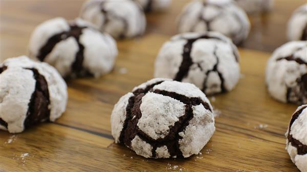 A batch of chocolate crinkle cookies dusted with powdered sugar sits on a wooden surface. The cookies have a distinctive cracked appearance, with dark chocolate showing through the white coating.