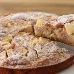 A close-up of a freshly baked apple cake dusted with powdered sugar. A slice is being lifted out, revealing the moist, apple-packed interior. The cake sits on a white plate with a wooden surface in the background.