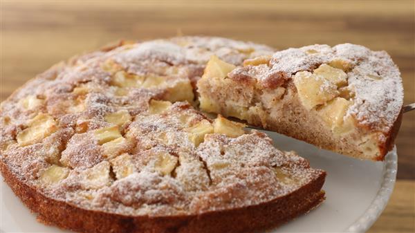 A close-up of a freshly baked apple cake dusted with powdered sugar. A slice is being lifted out, revealing the moist, apple-packed interior. The cake sits on a white plate with a wooden surface in the background.