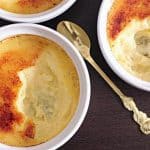 Three bowls of crème brûlée are shown on a dark surface, all with a caramelized sugar topping. One bowl has a spoon with a bite taken out. The golden spoons, each with a decorative handle, rest beside or in the bowls.