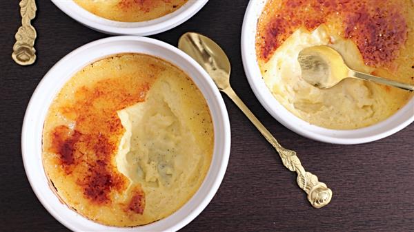 Three bowls of crème brûlée are shown on a dark surface, all with a caramelized sugar topping. One bowl has a spoon with a bite taken out. The golden spoons, each with a decorative handle, rest beside or in the bowls.