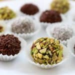 A variety of round truffles are arranged on a white plate. Each truffle is topped with different coatings, such as chocolate sprinkles, chopped pistachios, and powdered sugar. They are placed in small white paper cups.