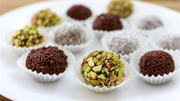 A variety of round truffles are arranged on a white plate. Each truffle is topped with different coatings, such as chocolate sprinkles, chopped pistachios, and powdered sugar. They are placed in small white paper cups.