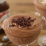 Two glass dishes filled with chocolate mousse, garnished with chocolate sprinkles, sit on a wooden table. In the background, there is a jar of Nutella and two spoons. The mousse has a creamy, smooth texture.