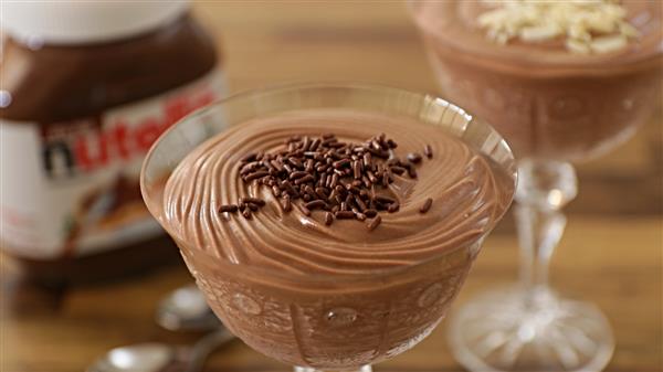 Two glass dishes filled with chocolate mousse, garnished with chocolate sprinkles, sit on a wooden table. In the background, there is a jar of Nutella and two spoons. The mousse has a creamy, smooth texture.