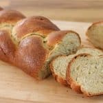 A loaf of braided bread sits on a wooden cutting board. Part of the loaf has been sliced, and several slices are shown in the foreground, revealing a slightly green-tinted, airy interior. The crust is golden brown.