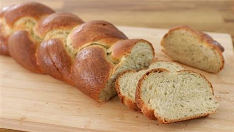 A loaf of braided bread sits on a wooden cutting board. Part of the loaf has been sliced, and several slices are shown in the foreground, revealing a slightly green-tinted, airy interior. The crust is golden brown.