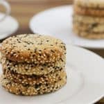 A stack of four sesame seed cookies rests on a white plate. In the background, another plate with more stacked cookies and a white cup are slightly blurred on a wooden surface.