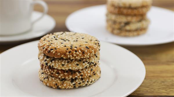 A stack of four sesame seed cookies rests on a white plate. In the background, another plate with more stacked cookies and a white cup are slightly blurred on a wooden surface.