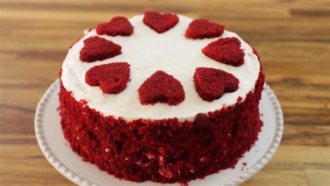 A red velvet cake with white frosting, topped with eight heart-shaped pieces of red velvet cake arranged in a circle on top. The sides are coated with red velvet cake crumbs, and the cake is placed on a white, round plate. The background is a wooden surface.