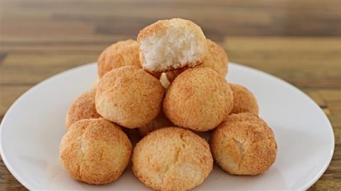 A white plate with a pyramid of golden-brown coconut macaroons. The macaroon on the top has a bite taken out, revealing a fluffy interior. The background is a wooden table.