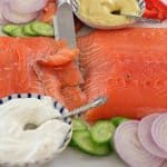 A close-up of a fresh, sliced salmon fillet surrounded by sliced cucumbers, red onions, tomatoes, and red bell peppers. Two bowls containing white and yellow sauces with spoons are also visible. A knife rests on the salmon.