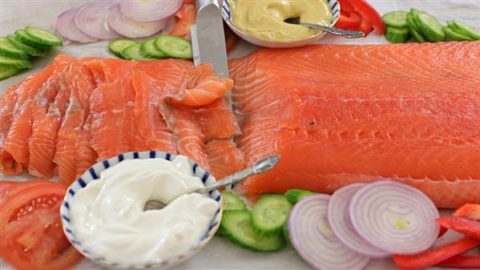 A close-up of a fresh, sliced salmon fillet surrounded by sliced cucumbers, red onions, tomatoes, and red bell peppers. Two bowls containing white and yellow sauces with spoons are also visible. A knife rests on the salmon.