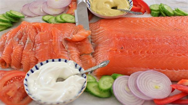 A close-up of a fresh, sliced salmon fillet surrounded by sliced cucumbers, red onions, tomatoes, and red bell peppers. Two bowls containing white and yellow sauces with spoons are also visible. A knife rests on the salmon.