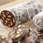 Two chocolate salami logs covered in powdered sugar are displayed on a wooden cutting board. One log is shown sliced, revealing the interior which contains visible chunks of nuts and biscuits. Several round slices are placed in front of the log.