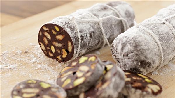 Two chocolate salami logs covered in powdered sugar are displayed on a wooden cutting board. One log is shown sliced, revealing the interior which contains visible chunks of nuts and biscuits. Several round slices are placed in front of the log.