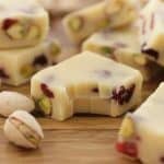 Close-up of several pieces of white chocolate fudge with visible pistachios and cranberries, arranged on a wooden surface. Scattered around the fudge are whole, shelled pistachios. One piece of fudge has a bite taken out of it.