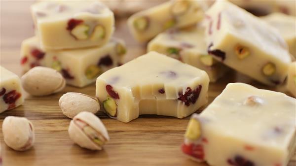 Close-up of several pieces of white chocolate fudge with visible pistachios and cranberries, arranged on a wooden surface. Scattered around the fudge are whole, shelled pistachios. One piece of fudge has a bite taken out of it.